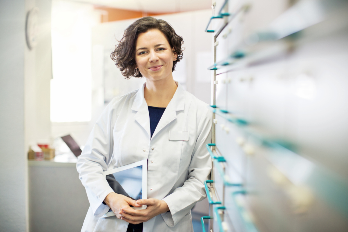 Portrait of confident young female pharmacist leaning to a medicine shelf with a digital tablet.