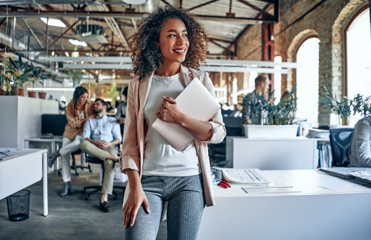 A woman walks through her office carrying a laptop while smiling.