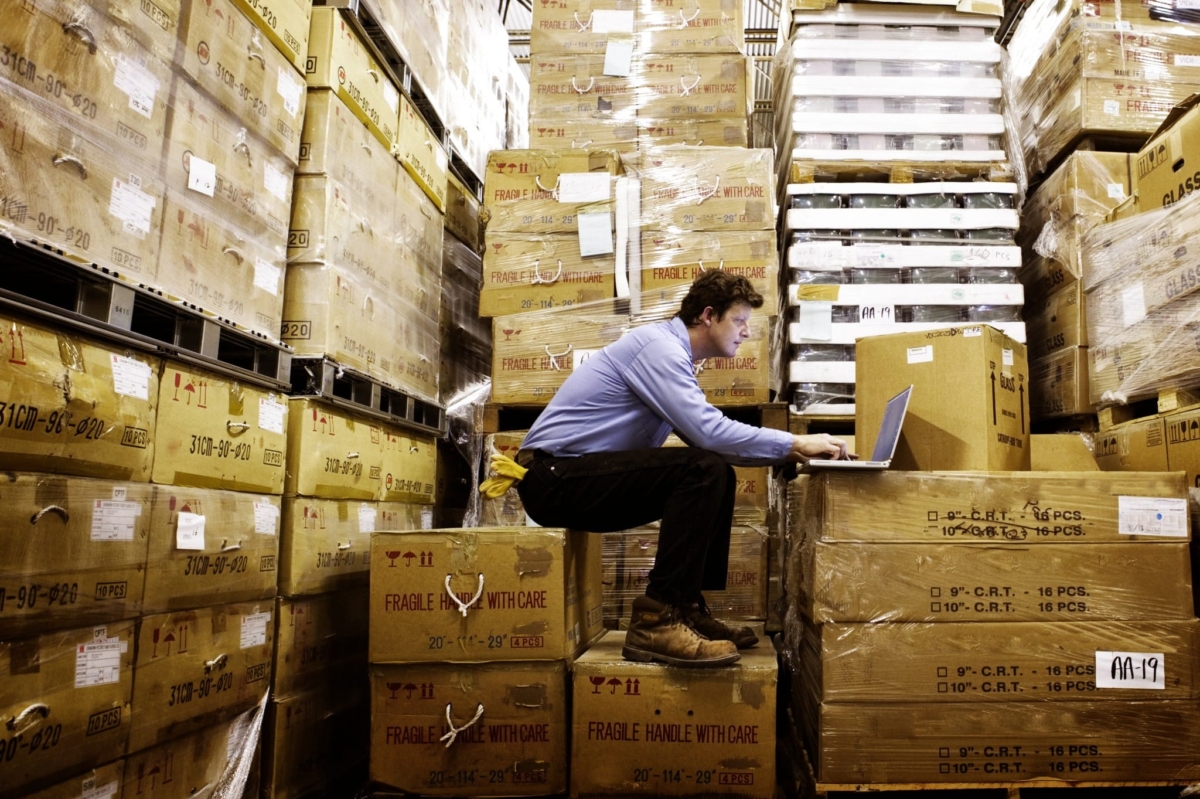 A man takes inventory while sitting on boxes in a warehouse.