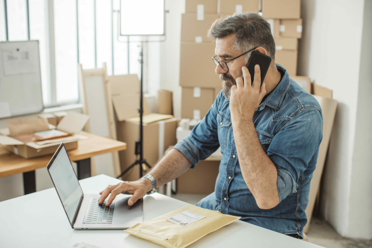 A man takes inventory on his laptop in a warehouse, while talking on the phone.