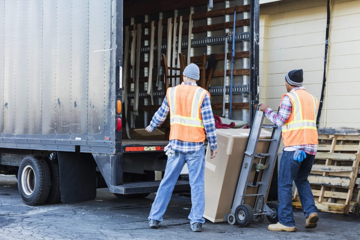 Two workers with a truck move a large box.